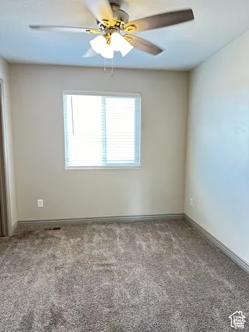 Neutral-toned interior featuring textured carpeting, a multi-blade ceiling fan with integrated light fixture, and a single-pane window with horizontal blinds