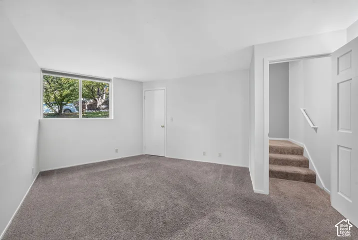 Neutral-toned carpeted flooring, large window, and interior door leading to carpeted stairway with a mounted handrail