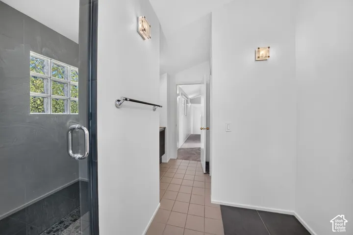 Walk-in shower featuring gray tiled walls, a glass door, a privacy block window, and square-tile flooring, accented by two mounted wall sconces