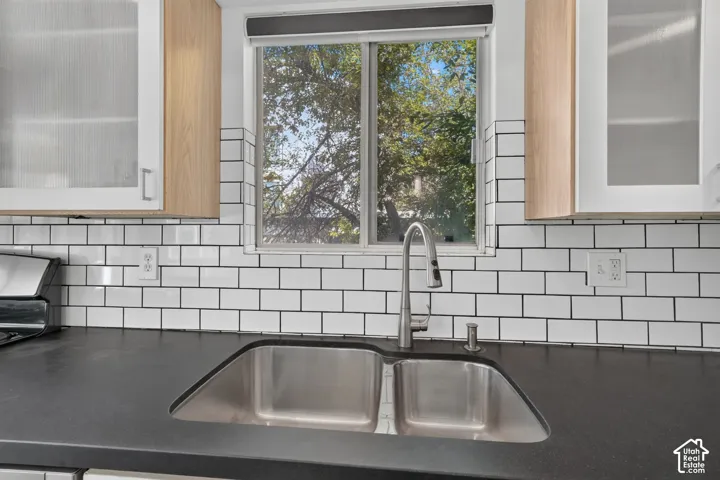 Double-basin stainless steel sink with a high-arch pull-down faucet, dark solid-surface countertop, white subway tile backsplash, and light-tone wood upper cabinetry with frosted glass inserts