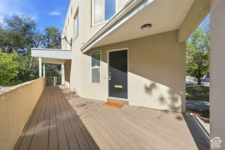 Covered balcony featuring wood-finish decking, stucco exterior walls, solid-core entry door with mail slot, and flush-mount exterior lighting