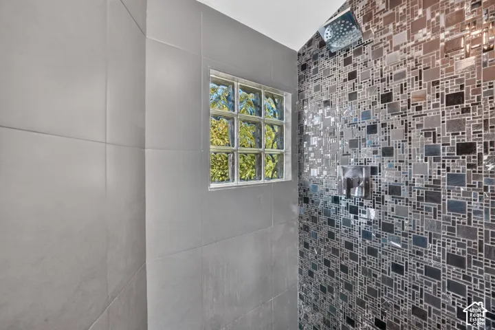 Shower stall featuring large-format grey wall tiles, a metallic mosaic-tiled accent wall, a square glass block window, and a wall-mounted rainfall shower head