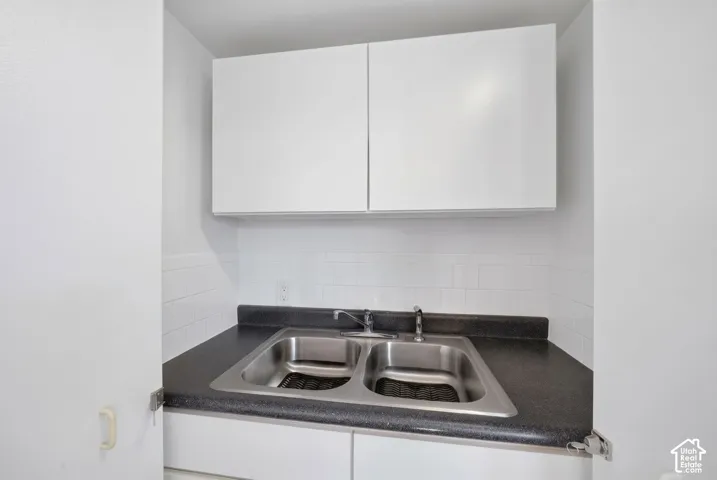 Kitchen area featuring white cabinetry, a dark solid-surface countertop, a dual-basin stainless steel sink, and a white tile backsplash