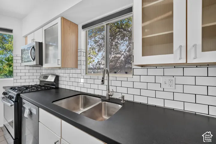 Kitchen featuring white cabinetry with glass-panel inserts, solid-surface countertops, dual-basin stainless steel sink with high-arc faucet, and subway tile backsplash