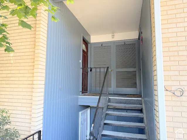 Exterior entry featuring tan brick masonry, vertical blue siding, a dark-toned door with a glass insert, and a metal staircase railing