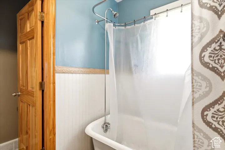Clawfoot tub featuring an exposed metal shower riser, beadboard wainscoting with decorative trim, and natural wood-finish door
