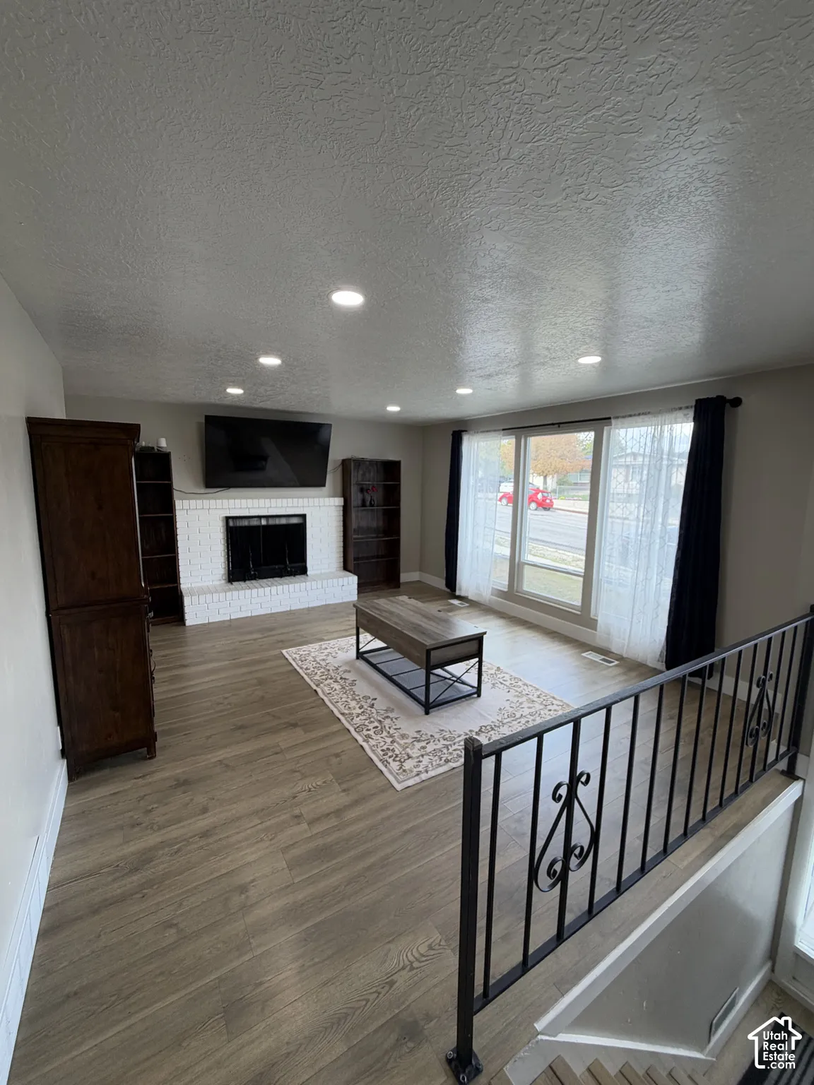 Living area featuring wood-finish flooring, white brick fireplace, recessed lighting, and metal stair railing