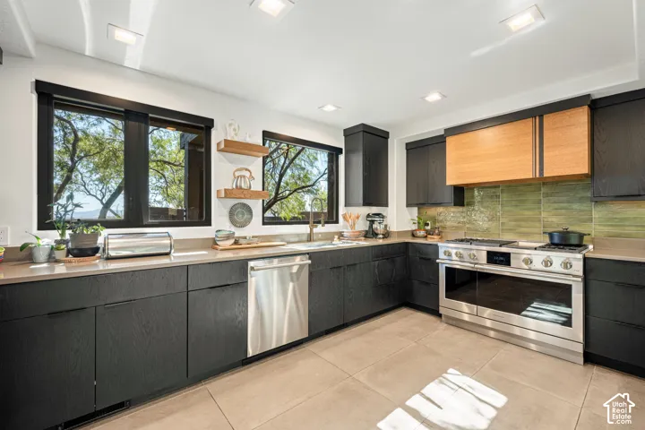 Kitchen featuring flat-panel cabinetry in matte black and wood finishes, stainless steel appliances, a green tile backsplash, and quartz countertops