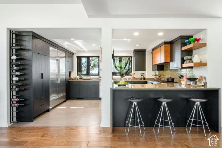 Kitchen featuring matte black cabinetry, stainless steel professional-grade refrigerator, waterfall-edge countertop, built-in wine rack, and natural wood floating shelving