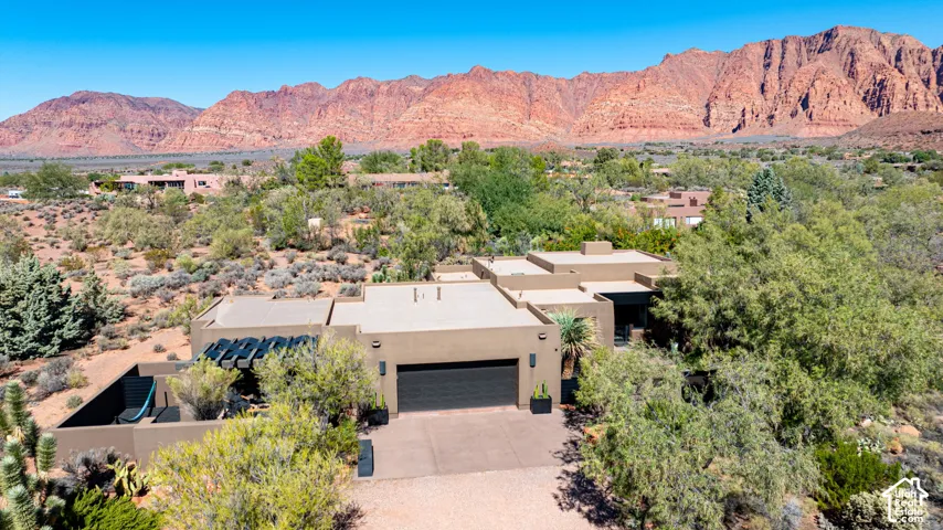 Single-story stucco residence featuring a flat roofline, attached multi-car garage, gated courtyard, and desert landscape set against a red mountain backdrop