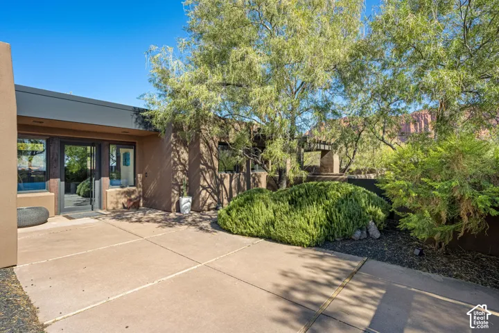 Stucco exterior facade featuring a concrete patio, wood-beam pergola, and mature landscaping with rock ground cover