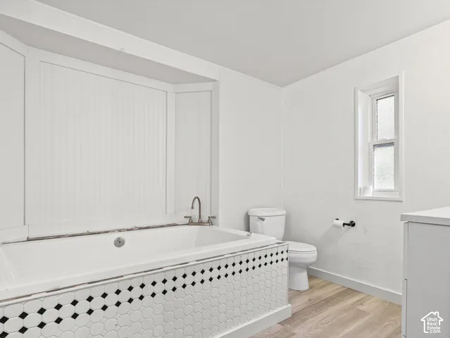 Deep huge soaking tub with beadboard wall paneling, geometric tile apron, matte black faucet fixtures, and wood-finish flooring