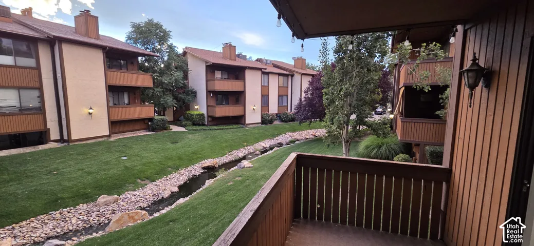 Covered balcony featuring wood-slat railings and wall-mounted light fixtures overlooking a landscaped common area with a stone-lined water feature and manicured lawn