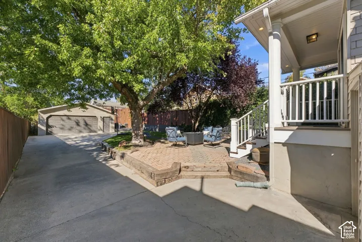 Rear exterior space featuring a concrete driveway, paver patio, wood timber retaining wall, covered porch with column supports, and a detached garage
