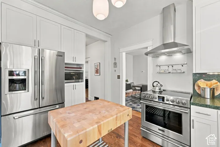 Kitchen featuring white shaker-style cabinetry, stainless steel appliances, a professional-grade range hood, dark solid-surface countertops, and wood-finish flooring
