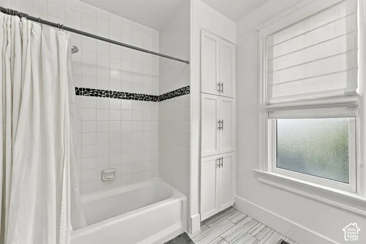Bathroom featuring a white porcelain tub, floor-to-ceiling white cabinetry with brushed-finish hardware, tile wall surrounds with a dark stone-textured accent border, and tile flooring