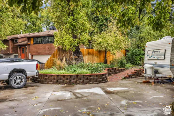 Concrete driveway area featuring a tiered retaining wall, brick paver path, wood-toned exterior siding, and mature tree landscaping