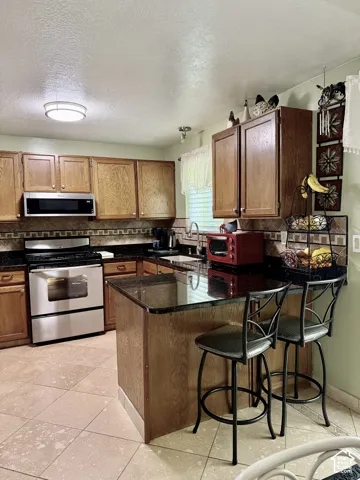 Kitchen featuring wood cabinetry, dark stone-finish countertops, stainless steel appliances, a tile backsplash with decorative accents, and tiled flooring