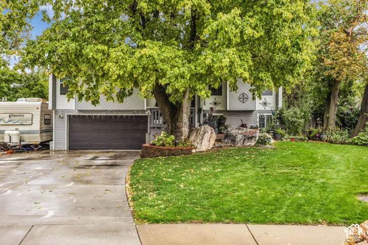 Split-level exterior featuring light-toned vertical siding, an attached two-car garage, concrete driveway, stone-accented landscaping, and mature foliage