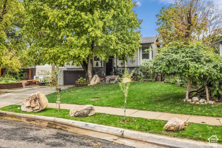 Split-level exterior featuring light-toned siding, attached garage, mature trees, ornamental landscaping, and natural boulder accents
