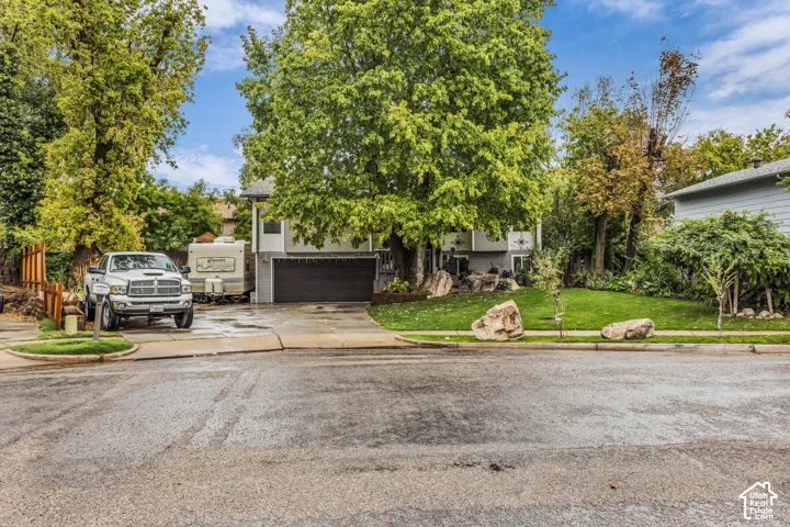 Exterior view of residence featuring a multi-level structure with an attached garage, paved driveway, mature trees, and landscaping accented by large decorative boulders