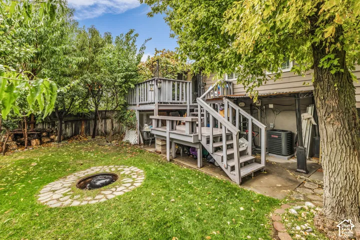 Multi-level wooden deck featuring stairs, stone-bordered fire pit, manicured lawn, and mature trees