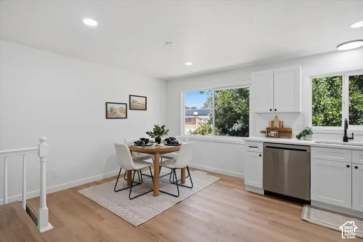 White shaker-style cabinetry, stainless steel dishwasher, wood-finish flooring, recessed lighting, and large windows providing natural light