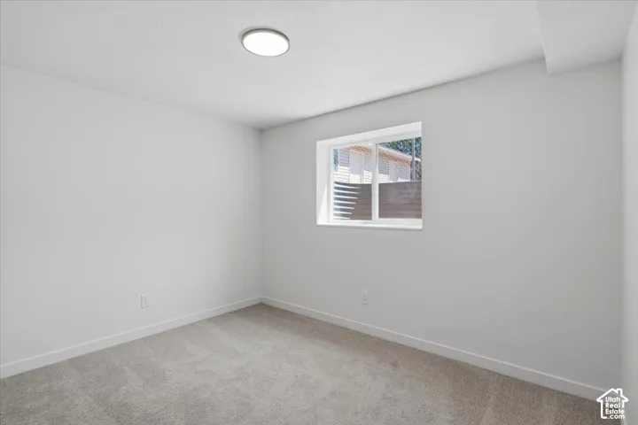 Neutral-toned room featuring neutral carpeting, white walls, flush-mount ceiling lighting, and a sliding window with an egress well