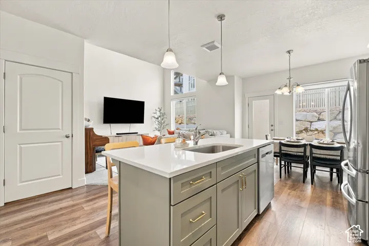 Kitchen island with quartz countertop, undermount stainless steel sink, sage green cabinetry with brass-finish hardware, wood-finish flooring, and pendant lighting