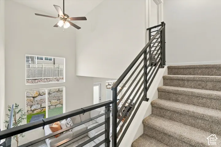 Staircase featuring carpeted steps and a custom metal horizontal-rail banister, soaring vaulted ceilings, a multi-blade ceiling fan, and stacked windows