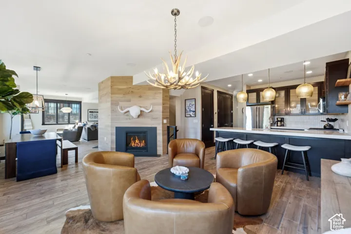 Open floor plan featuring wood-finish flooring, central fireplace with wood-panel surround, tiered kitchen island with pendant lighting, and decorative antler-style chandelier