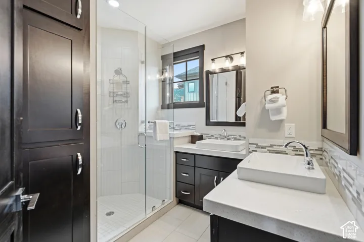 Bathroom featuring a dual-sink vanity with light-colored countertops, square vessel sinks, dark-toned cabinetry, and a walk-in glass-enclosed shower with tile surround