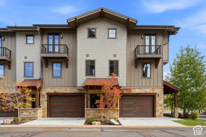 Multi-level exterior featuring vertical siding, stone masonry, metal roofing accents, private balconies with metal railings, and dual two-car garages with wood-finish sectional doors