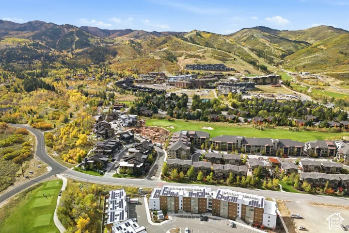 Aerial perspective of multi-story residential buildings featuring rooftop solar panel arrays, exterior stone and wood cladding, and paved surface parking, set against a backdrop of mountain slopes and mature foliage
