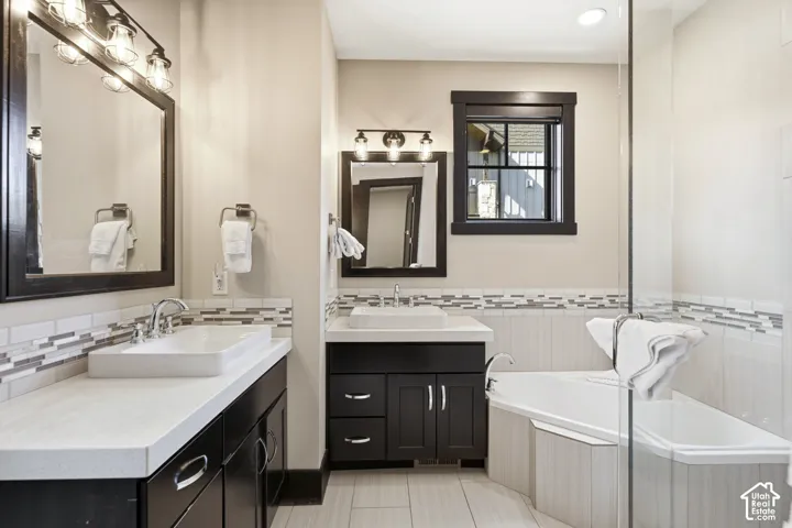 Dual-sink vanity with dark cabinetry, light-toned countertops, rectangular vessel sinks, tiled backsplash, and glass-enclosed soaking tub