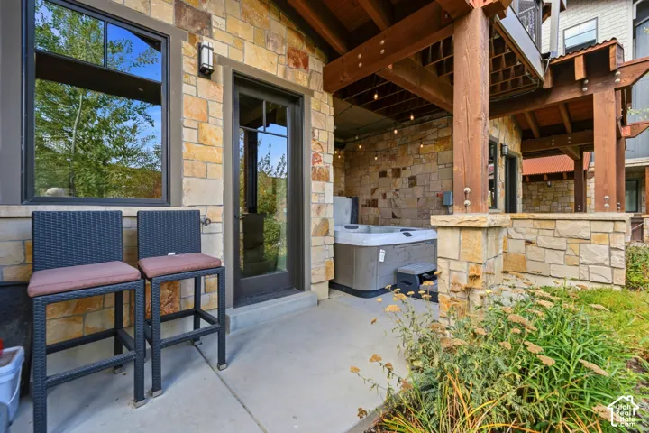Covered patio featuring stone exterior cladding, wood pergola support beams, concrete flooring, and built-in hot tub