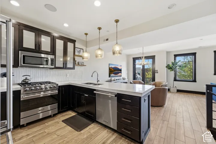 Kitchen featuring dark cabinetry with glass-panel upper doors, stainless steel appliances, a peninsula with stone-finish countertops, decorative pendant lighting, and wood-finish flooring