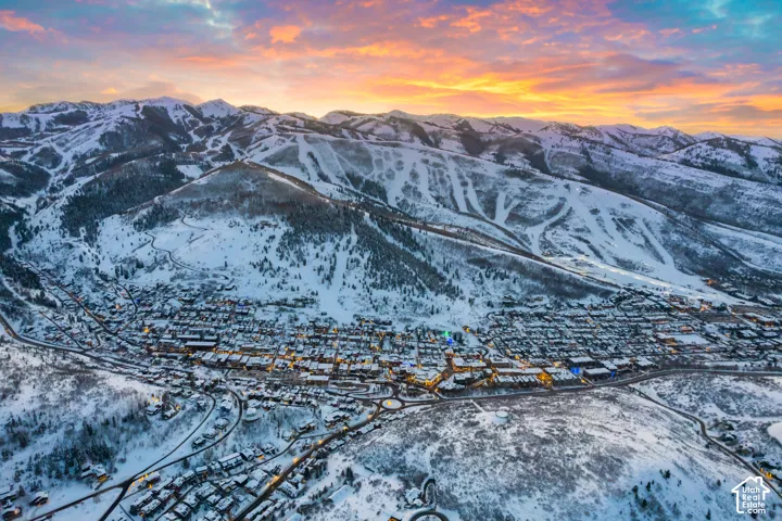 Aerial perspective featuring expansive snow-covered mountain terrain, illuminated valley infrastructure, and prominent ridgelines under a sunset sky