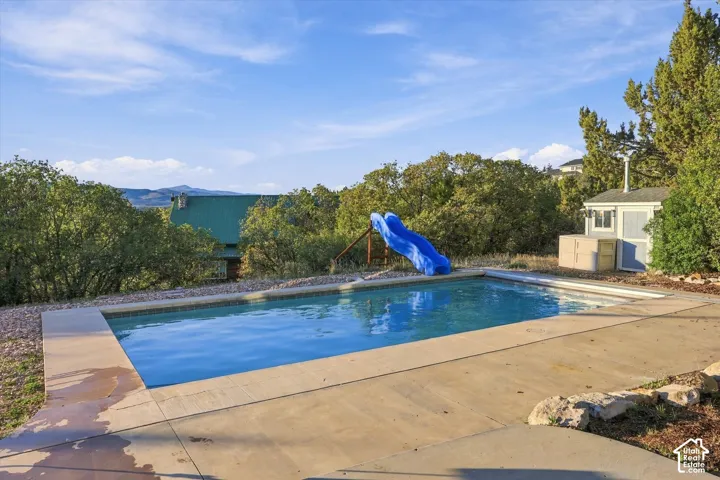 In-ground rectangular swimming pool with concrete decking, poolside water slide, and detached storage structure situated among mature trees