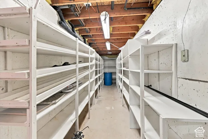 Storage area featuring extensive built-in white shelving, concrete wall construction, exposed wood joists, and dual linear light fixtures