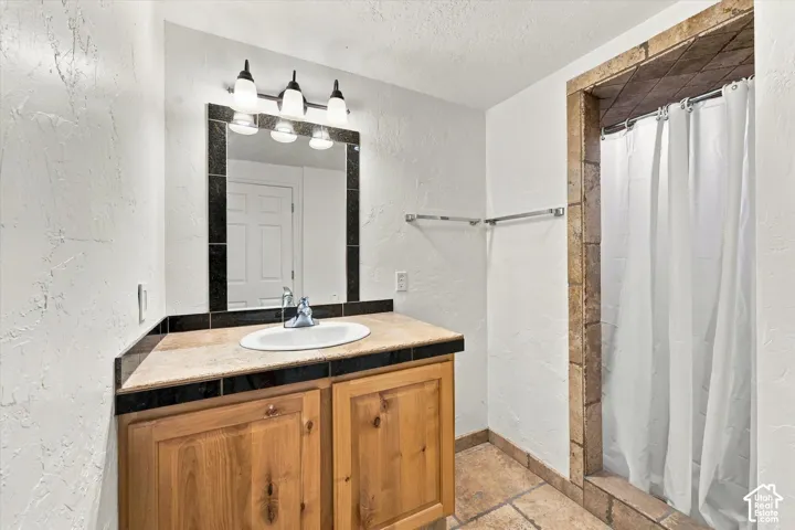 Basement Bathroom featuring a natural wood-finish vanity, stone-topped counter with integrated sink, three-light wall fixture, and tiled shower surround
