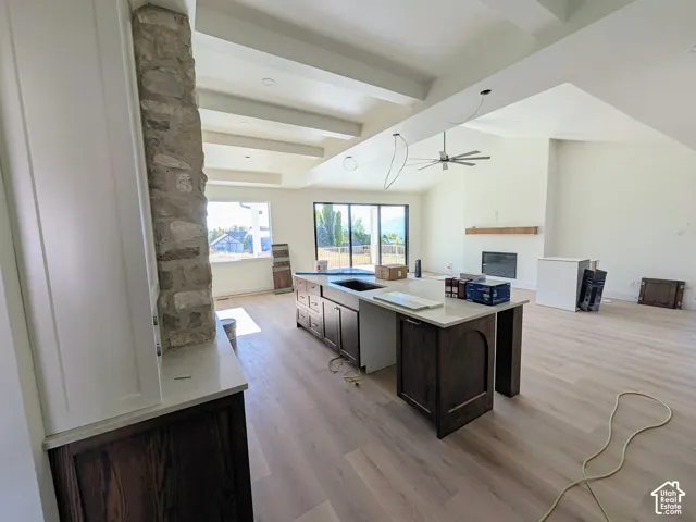 Open-concept area featuring wood-finish flooring, exposed ceiling beams, a central island with stone-finish countertops, a floor-to-ceiling stone-veneer column, and a fireplace with a wood mantel