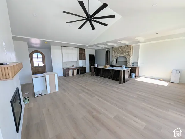 Vaulted ceilings featuring a multi-blade ceiling fan, wide-plank wood-finish flooring, central kitchen island with custom cabinetry, stone-surround fireplace, and arched wood-framed entry door