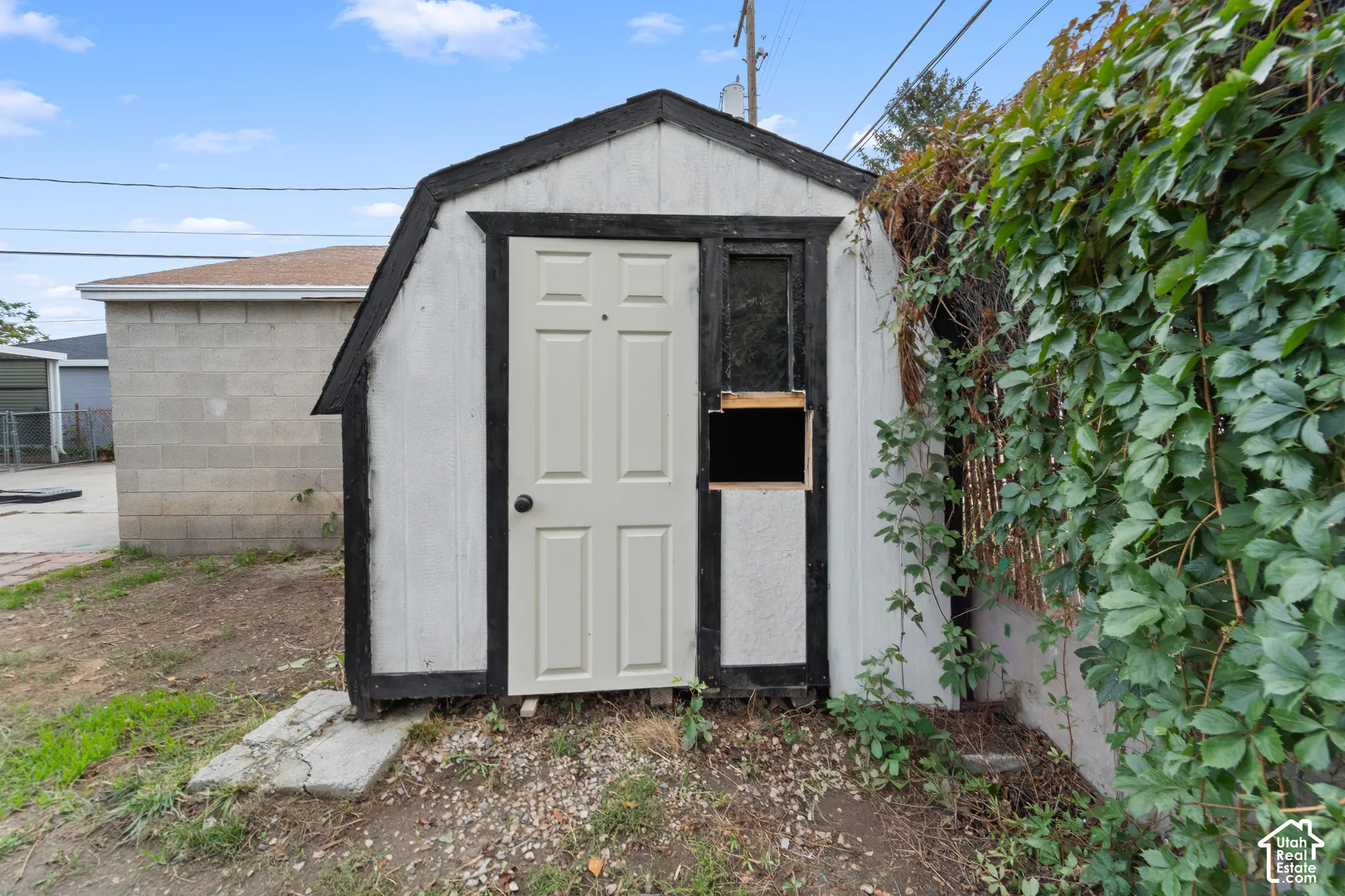 Exterior storage structure featuring a gambrel roofline, white paneling with contrasting dark trim, and a centered hinged door
