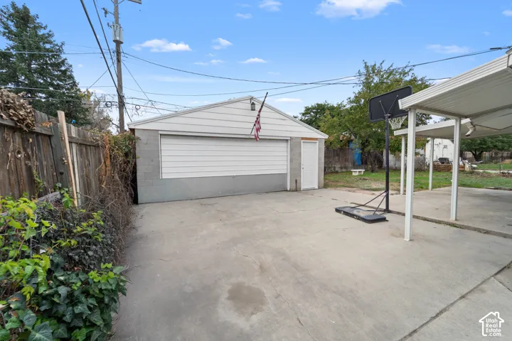 Detached garage featuring concrete block construction, a horizontal roll-up door, a dedicated pedestrian entry door, and an expansive concrete parking area covered by a metal-roofed carport