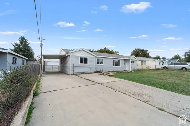 Single-story light grey siding exterior featuring a concrete driveway, covered carport, and metal chain-link fencing