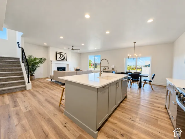 Open-concept interior featuring wood-finish flooring, a kitchen island with a neutral-tone countertop and under-mount sink, recessed lighting, a fireplace with a mantel, and staircase with a matte black metal railing