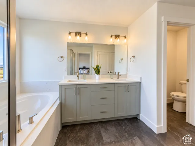 Dual-sink vanity with gray cabinetry and solid-surface countertop, soaking tub, polished metal hardware, and stone-finish tile flooring
