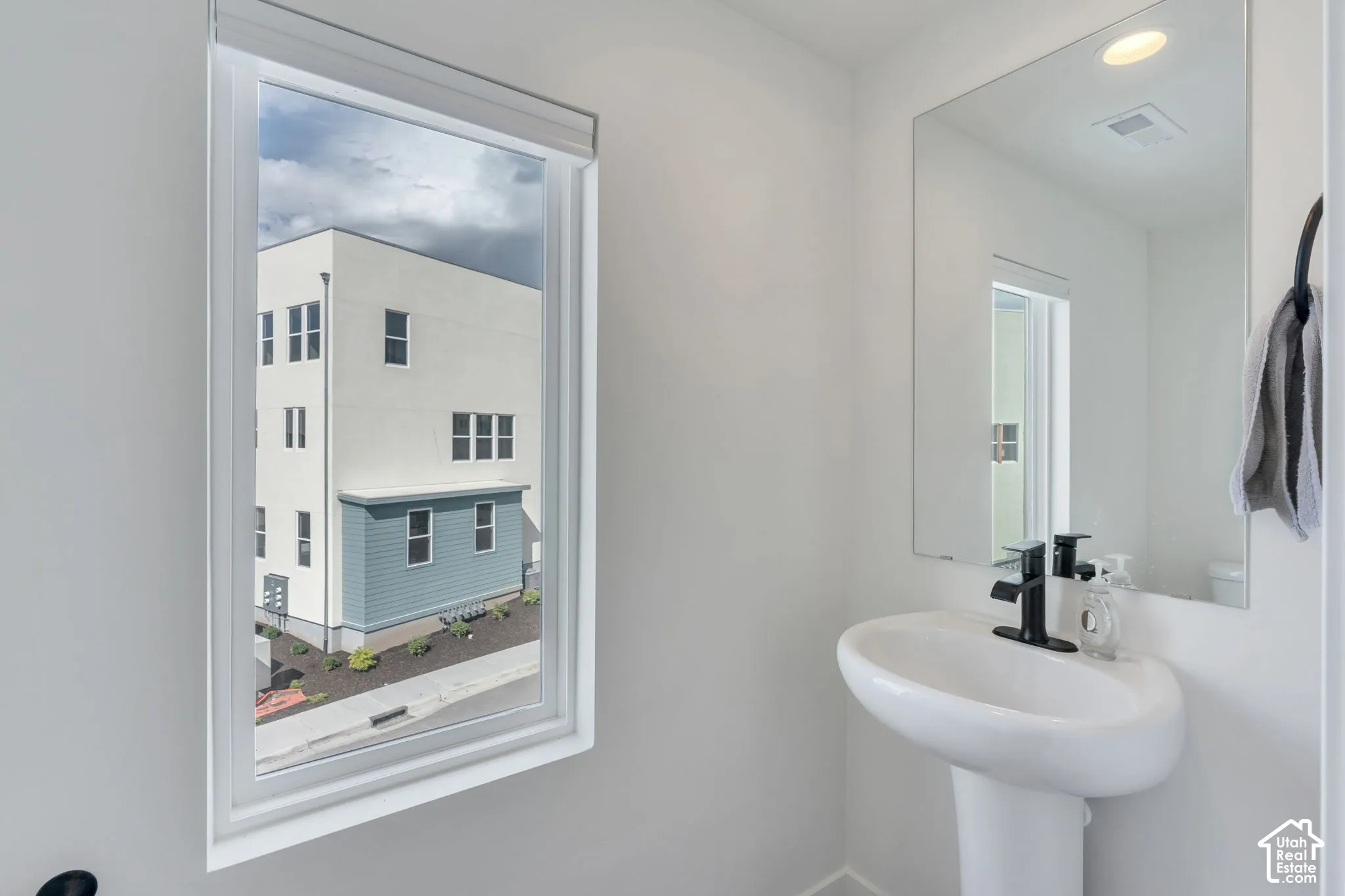 White pedestal sink featuring a matte black faucet, large frameless wall mirror, and window with adjustable roller shade