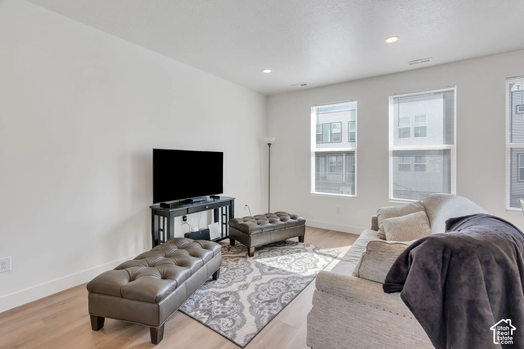 Living area featuring wood-finish flooring, neutral wall paint, and multiple large windows with integrated blinds, complemented by recessed ceiling lighting